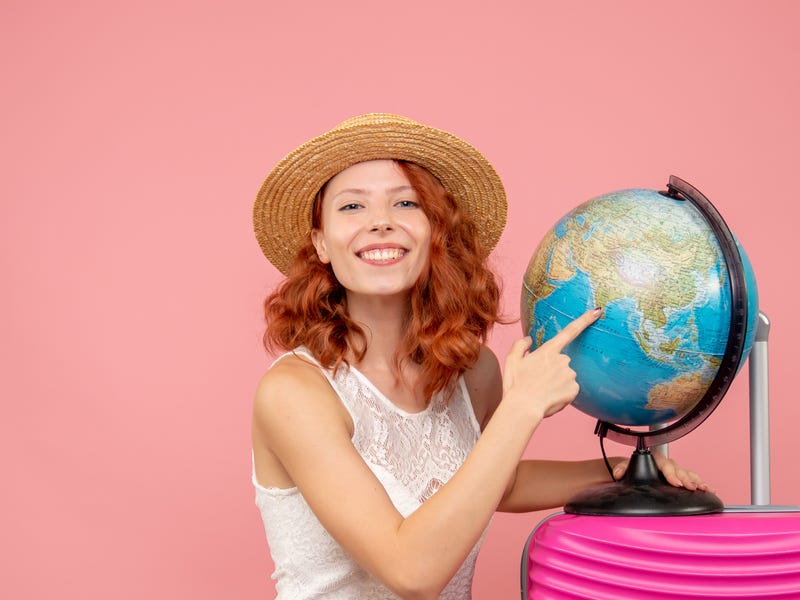 Mulher ao lado de um globo terrestre, com uma mala rosa pronta para viajar em inglês