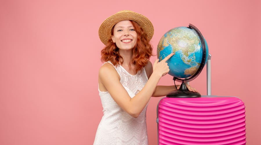 Mulher ao lado de um globo terrestre, com uma mala rosa pronta para viajar em inglês
