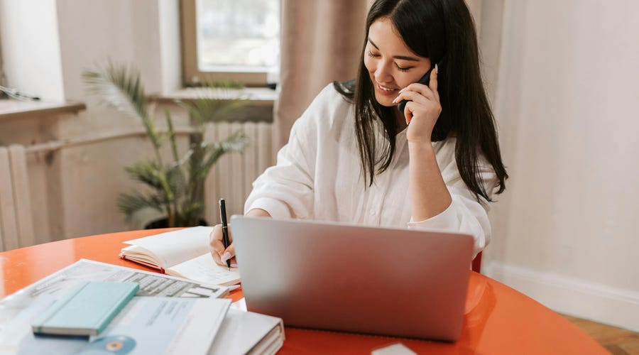 Mulher empreendedora em uma ligação anotando o que está escutando em um caderno enquanto está sentada em frente a um computador.