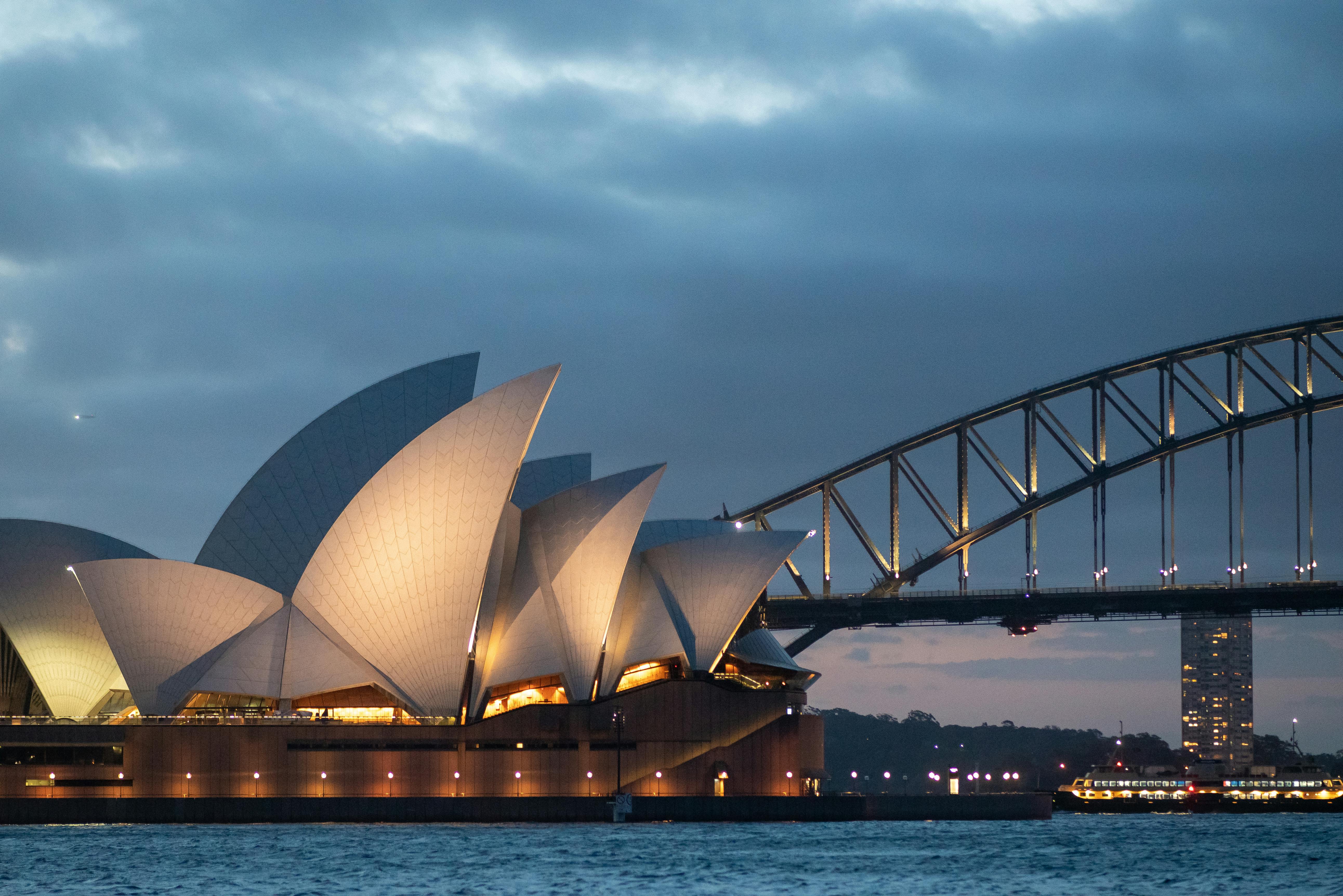 Imagem noturna da atração mais famosa em Sydney na Austrália, com uma ponte ao fundo.