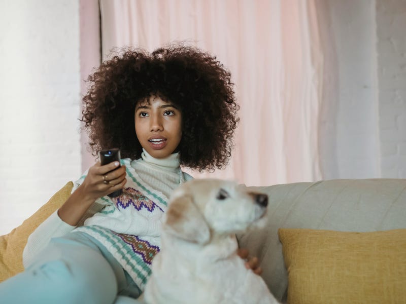 Jovem sentada no sofá com seu cachorro enquanto decide o que assistir na TV entre as séries em alemão que selecionou.