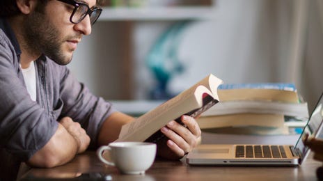 Homem utilizando óculos lendo um livro sentado em frente a uma mesa.