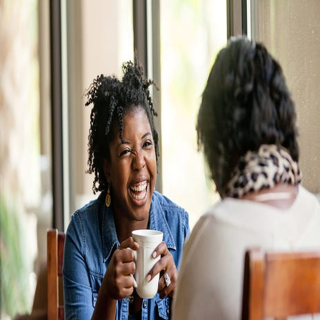 Mulher negra tomando um café com sua amiga, rindo das gírias alemãs que está aprendendo.