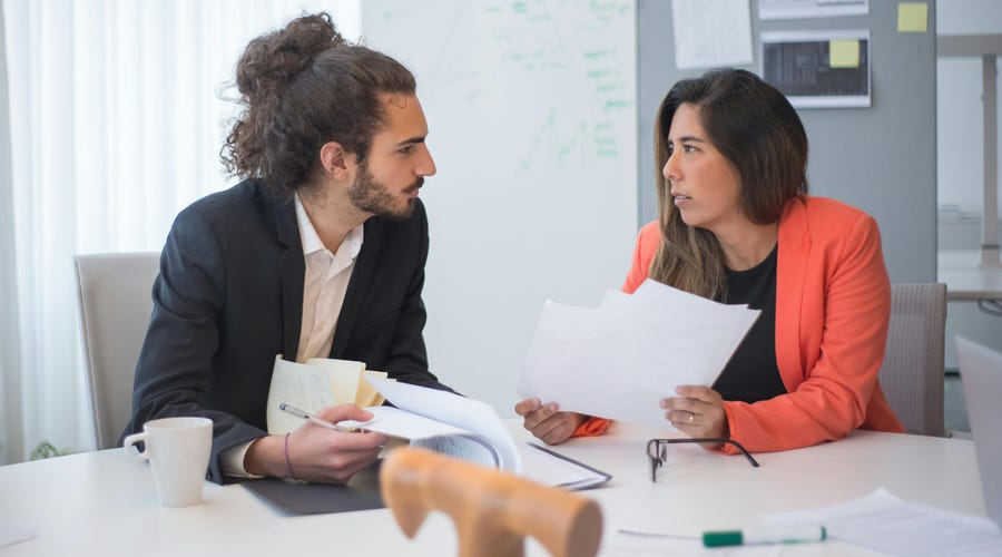 Mulher de cabelo comprido e homem com o cabelo cacheado preso em um coque durante uma de suas reuniões one on one.