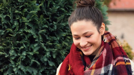 Mulher de cachecol e cabelo preso mexendo no celular em um banco de madeira ao ar livre.