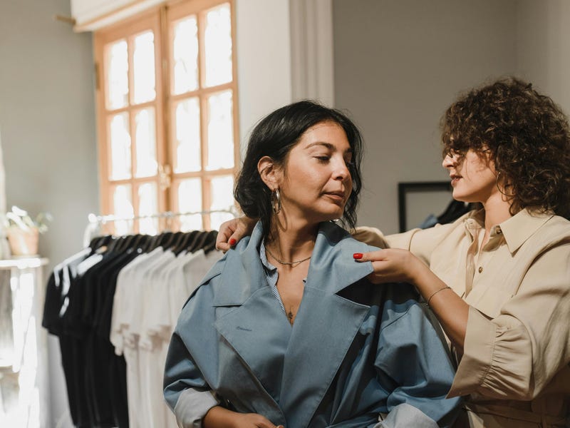 Mulher de casaco azul com botões em pé ao lado da mulher de casaco bege experimentando roupas em italiano.