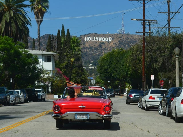 Carro vermelho andando pelas ruas de Los Angeles, nos Estados Unidos, com o letreiro de Hollywood no horizonte.