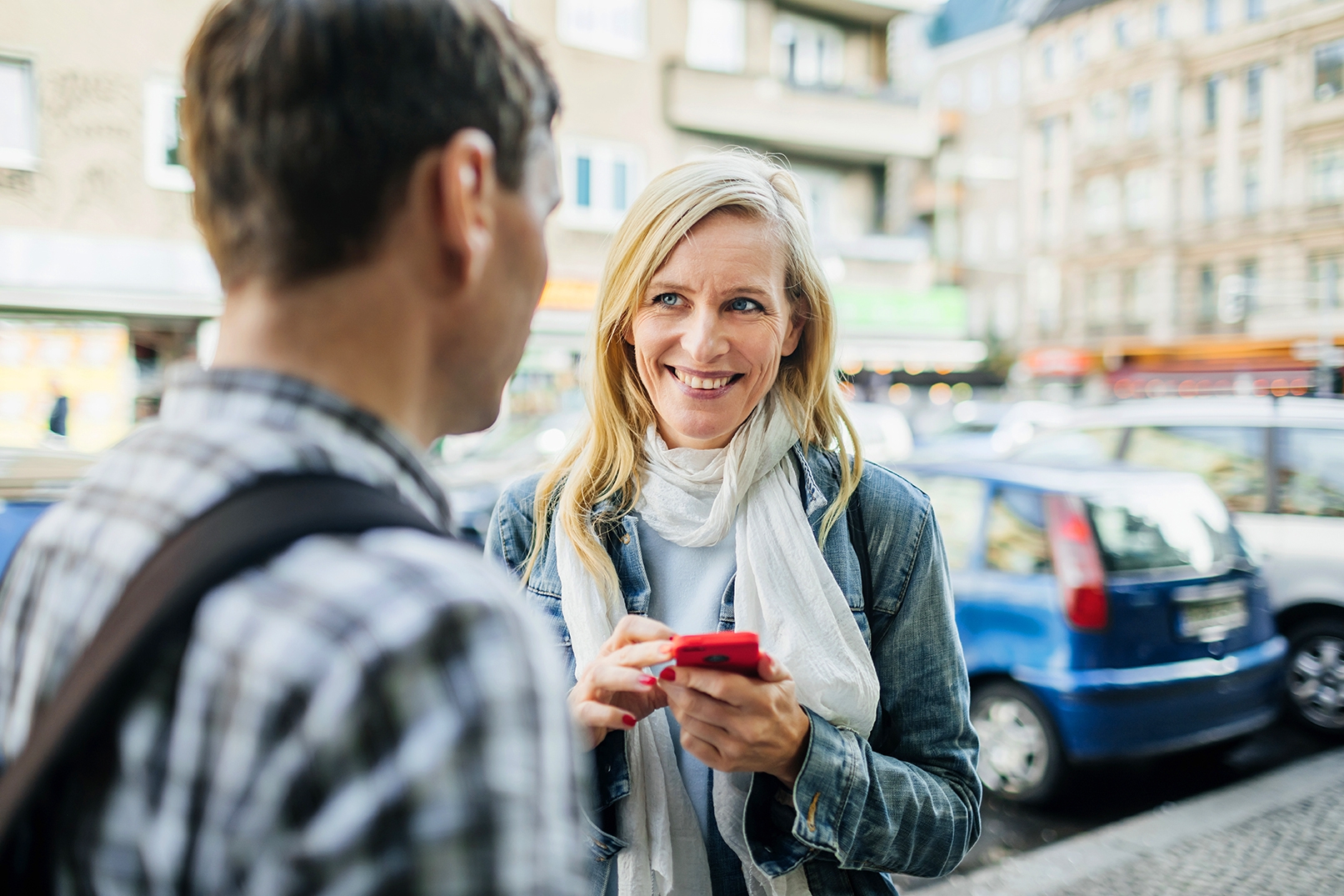 Mulher loira de olhos azuis segura seu celular vermelho enquanto sorri para estrangeiro com quem está conversando na rua.