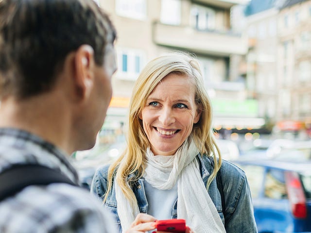 Mulher loira de olhos azuis segura seu celular vermelho enquanto sorri para estrangeiro com quem está conversando na rua.