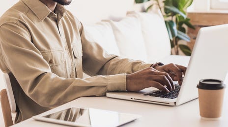 Homem jovem, vestindo uma camisa bege e óculos de grau, está sorrindo enquanto trabalha em seu notebook em casa.