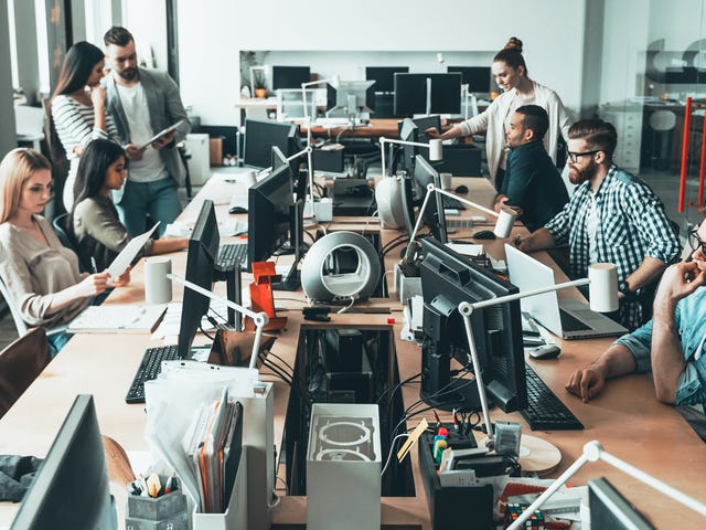 Mesa com diversos computadores e pessoas conversando e mexendo nestes computadores.