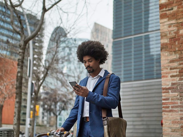 Homem negro parado em uma rua de Berlim, segurando sua bicicleta enquanto verifica sua nota do exame Telc b2 no celular.
