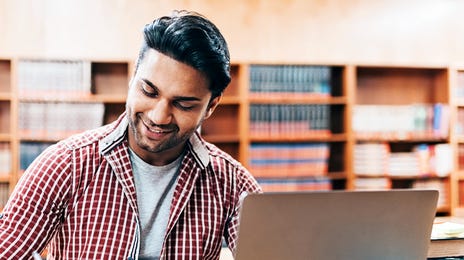 Homem em uma biblioteca fazendo anotações sobre algo que está lendo em seu laptop.