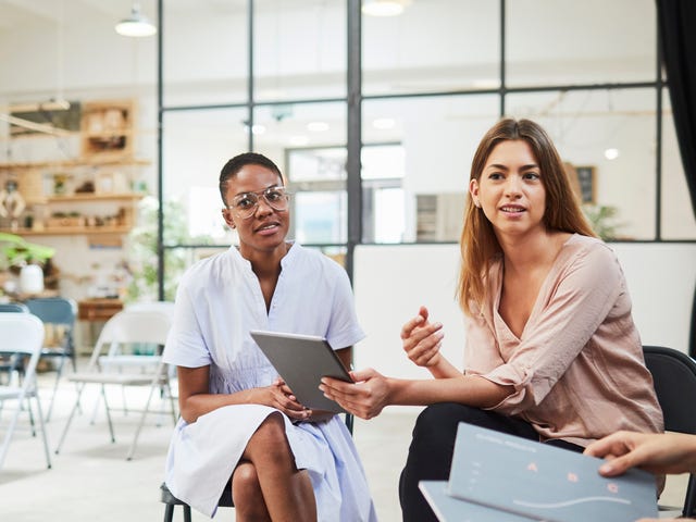 Duas jovens estudantes, uma negra de vestido e óculos, e uma branca de calça e camisa, sentadas em uma sala de preparação para o exame de proficiência TELC, conversando com colegas que realizarão a mesma prova.