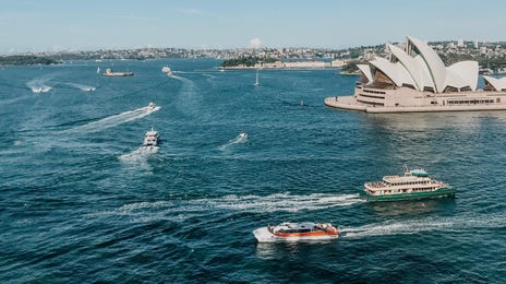 Vista aérea de Sydney, com mar azul e barcos navegando ao lado do ponto turístico mais visitado.