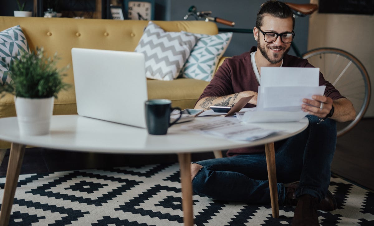 Homem de óculos sentado no chão com um laptop apoiado em sua mesa de centro enquanto revisa alguns papeis sorridente.