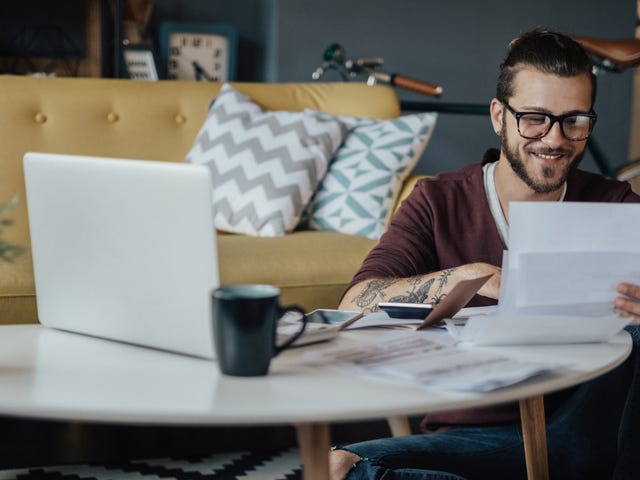 Homem de óculos sentado no chão com um laptop apoiado em sua mesa de centro enquanto revisa alguns papeis sorridente.