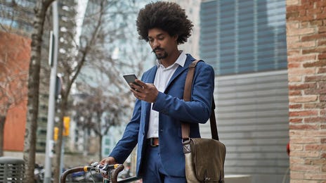 Homem negro parado em uma rua de Berlim, segurando sua bicicleta enquanto verifica sua nota do exame Telc b2 no celular.