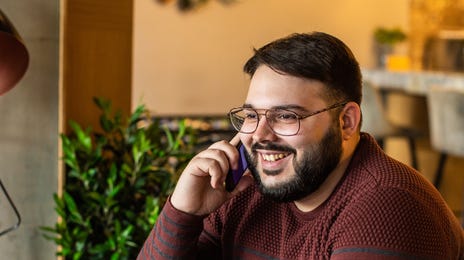 Homem falando ao telefone sorrindo enquanto mexe em um laptop.