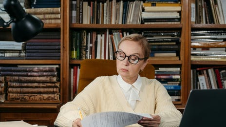 Mulher lendo um livro em uma mesa de escritório em frente a uma enorme estante de livros.