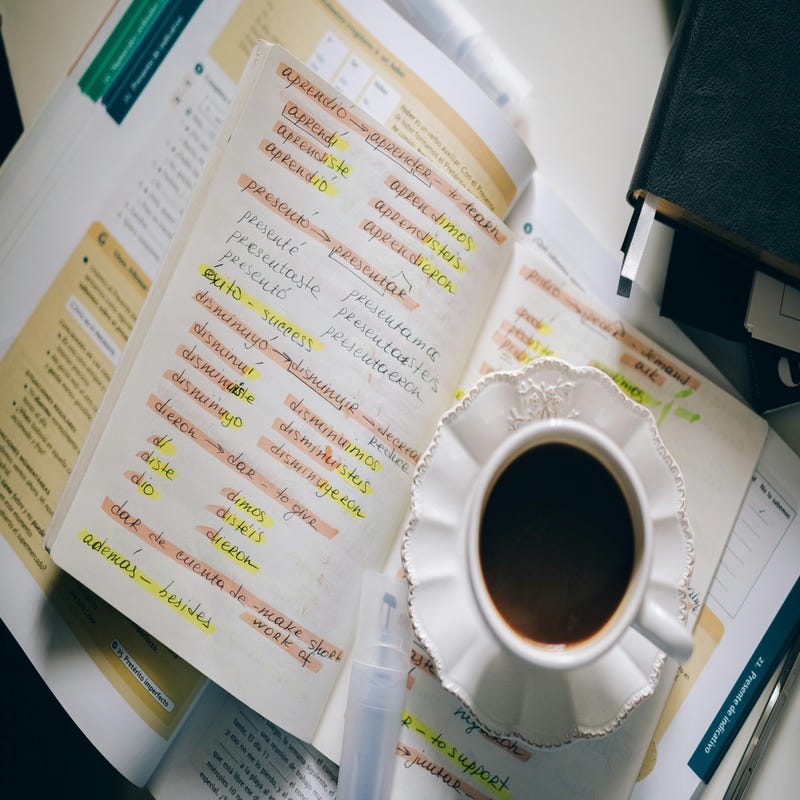 Livro e caderno de estudos com as palavras difíceis em espanhol grifadas, com uma xícara de café em cima.