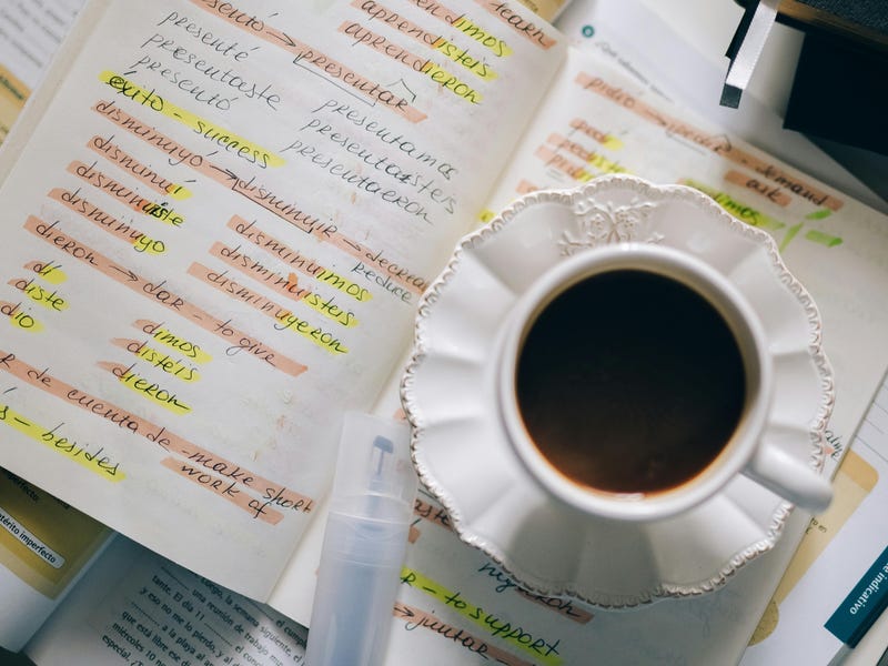 Livro e caderno de estudos com as palavras difíceis em espanhol grifadas, com uma xícara de café em cima.