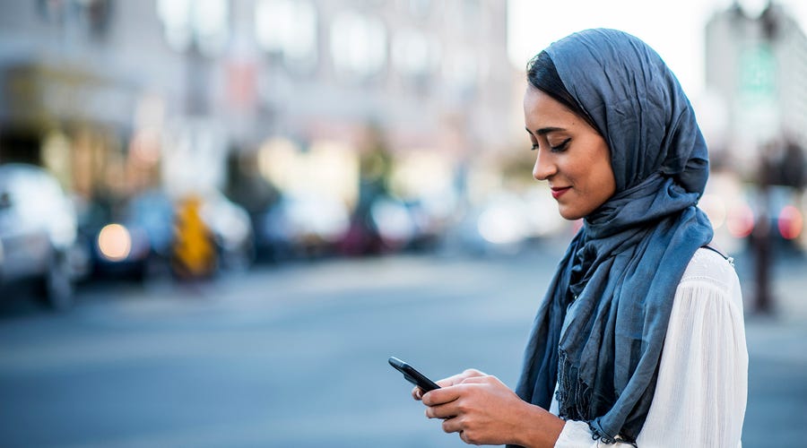 Mulher com jihab na cabeça está parada na calçada, espero seu ônibus enquanto checa o celular para saber as horas em francês.