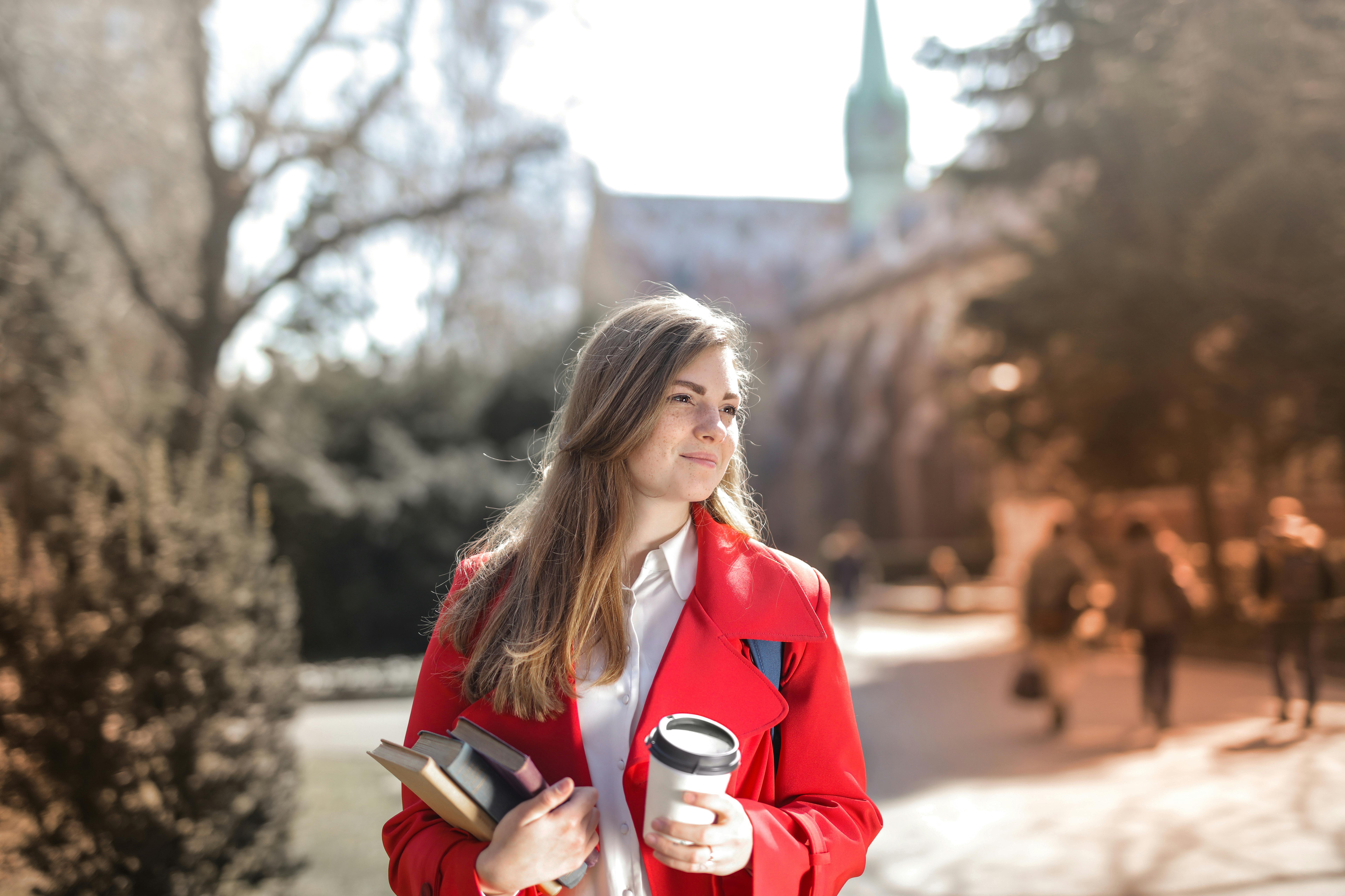 Mulher vestida de vermelho sorrindo em um país europeu, segurando livros e um café enquanto se prepara para a prova Telc Regular.