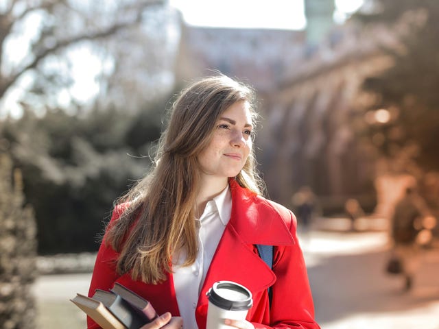 Mulher vestida de vermelho sorrindo em um país europeu, segurando livros e um café enquanto se prepara para a prova Telc Regular.