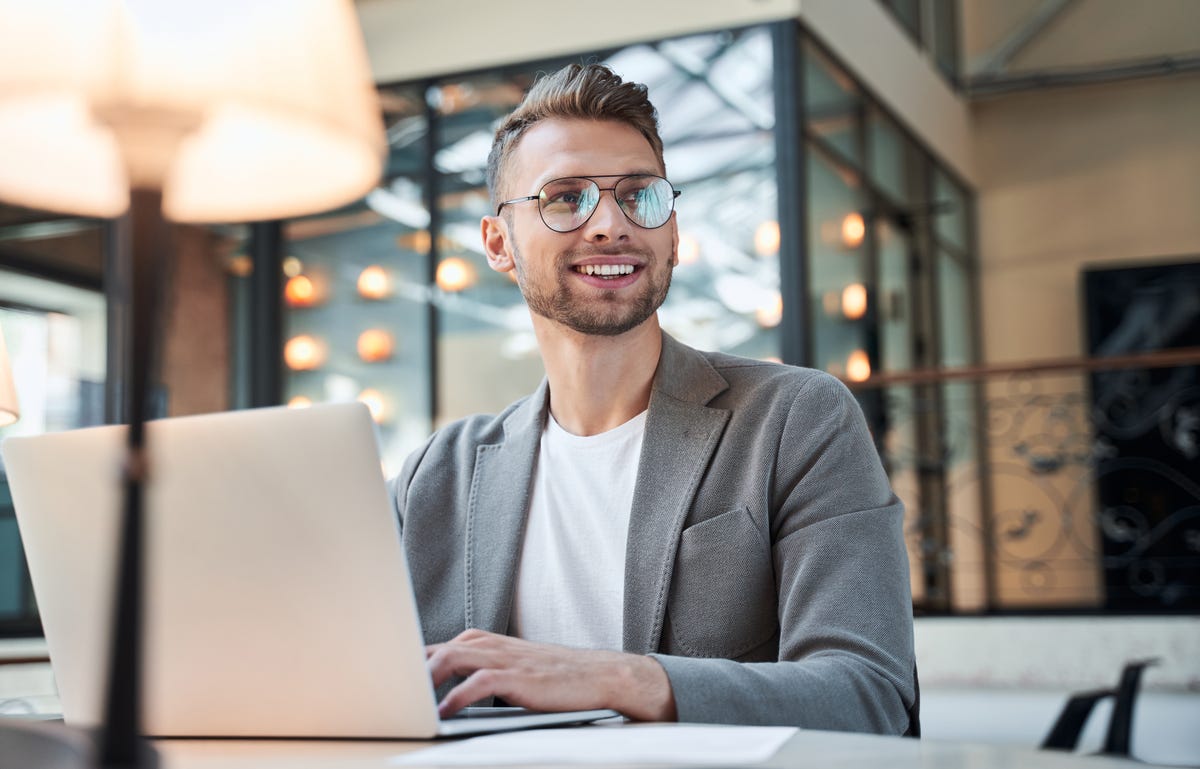 Homem loiro sorridente, vestindo um blazer cinza e um óculos de grau, está trabalhando com seu notebook em seu escritório.