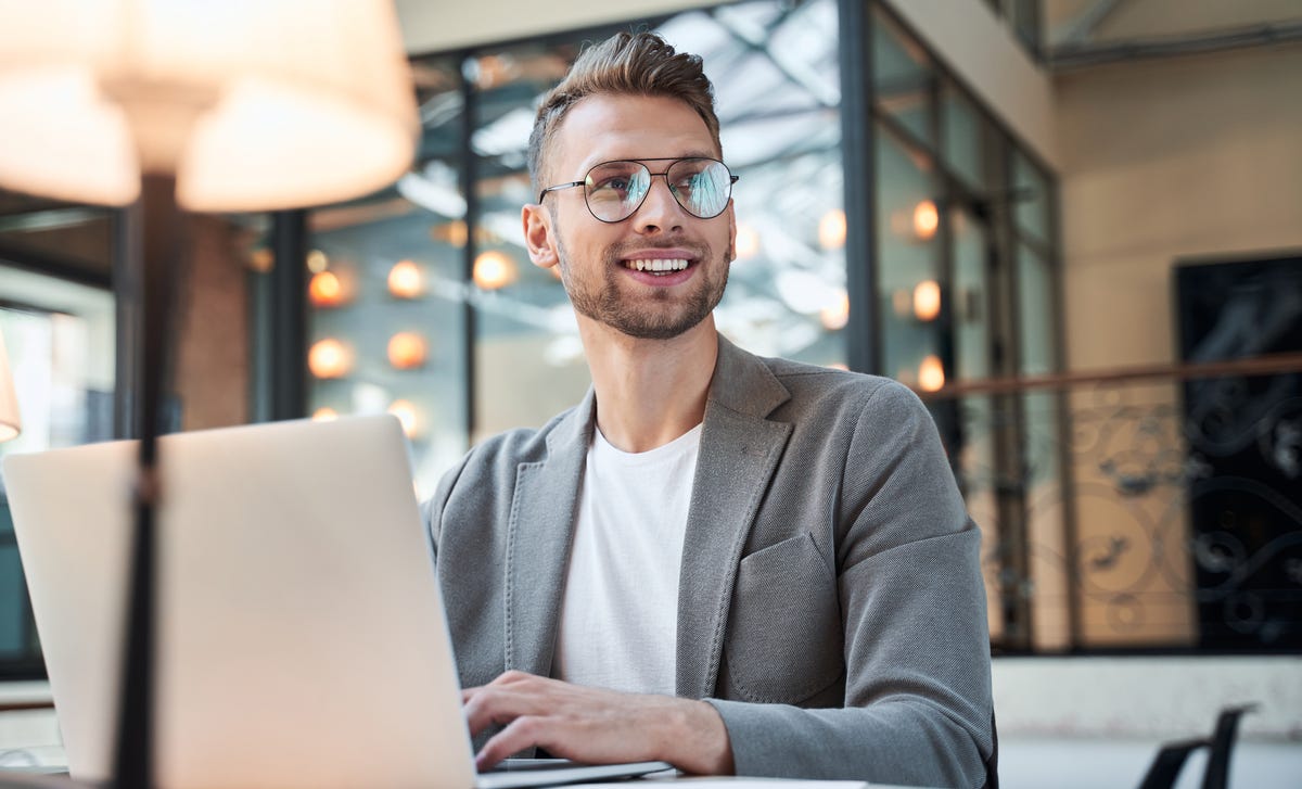 Homem loiro sorridente, vestindo um blazer cinza e um óculos de grau, está trabalhando com seu notebook em seu escritório.
