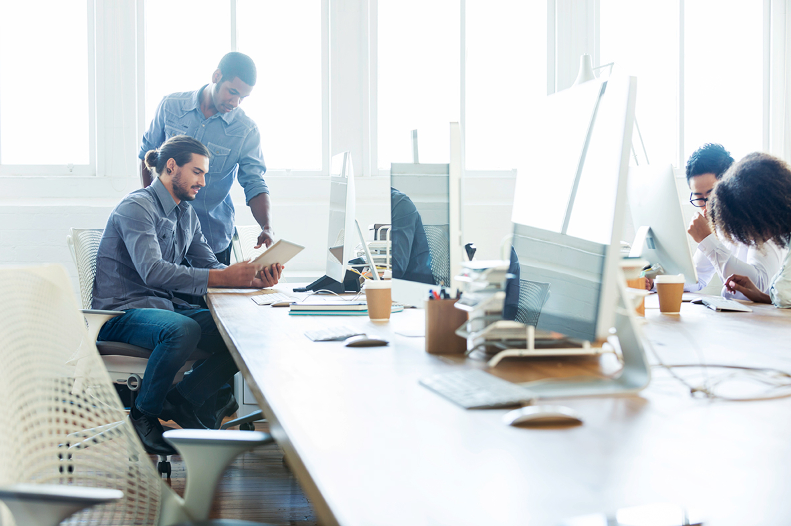 Escritório com mesas e computadores, onde dois funcionários comentam as novas diretrizes em inglês para TI.