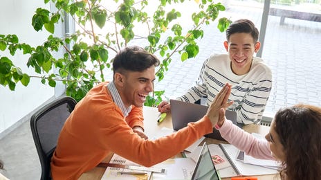 Students sitting around a table with their laptops and giving each other a high five during one of their group language classes