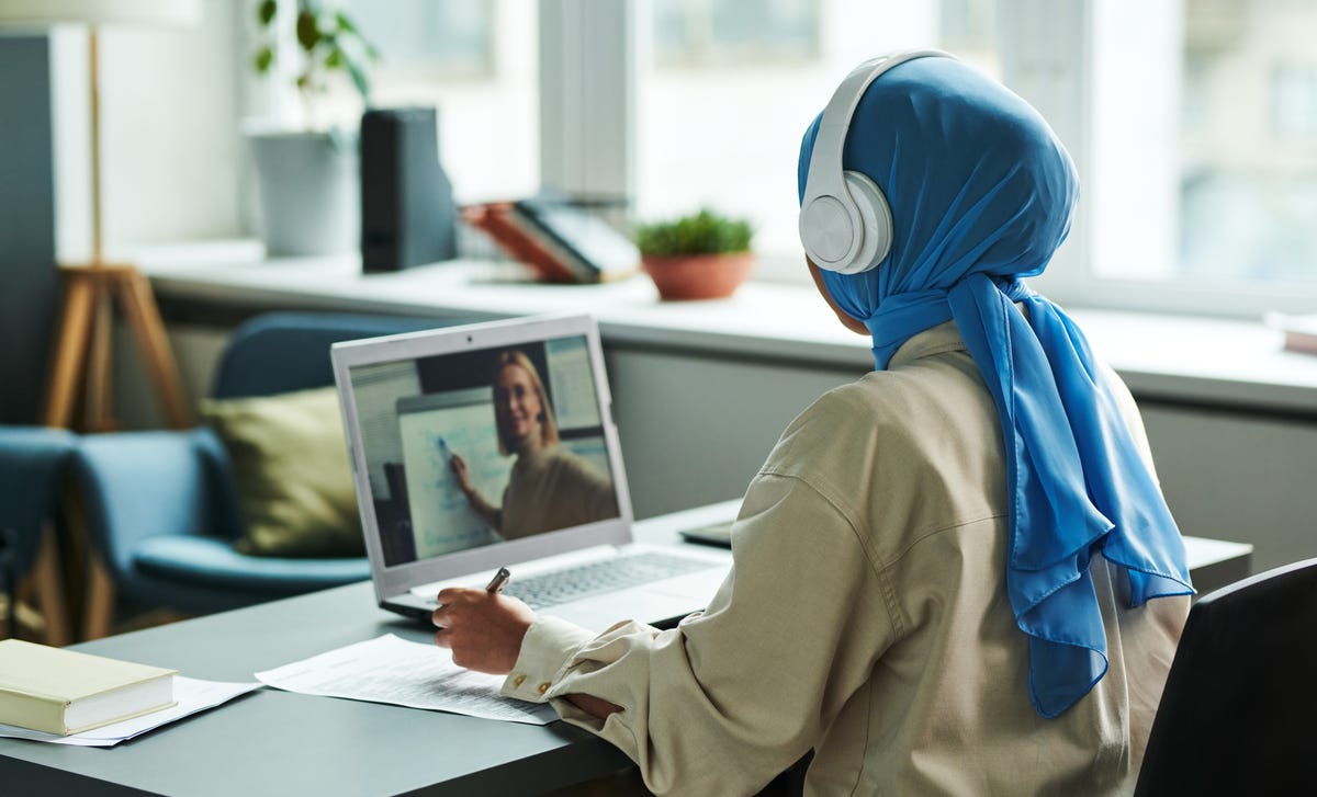Woman in a scarf wearing a headphone and attending one-on-one language tutoring from her laptop