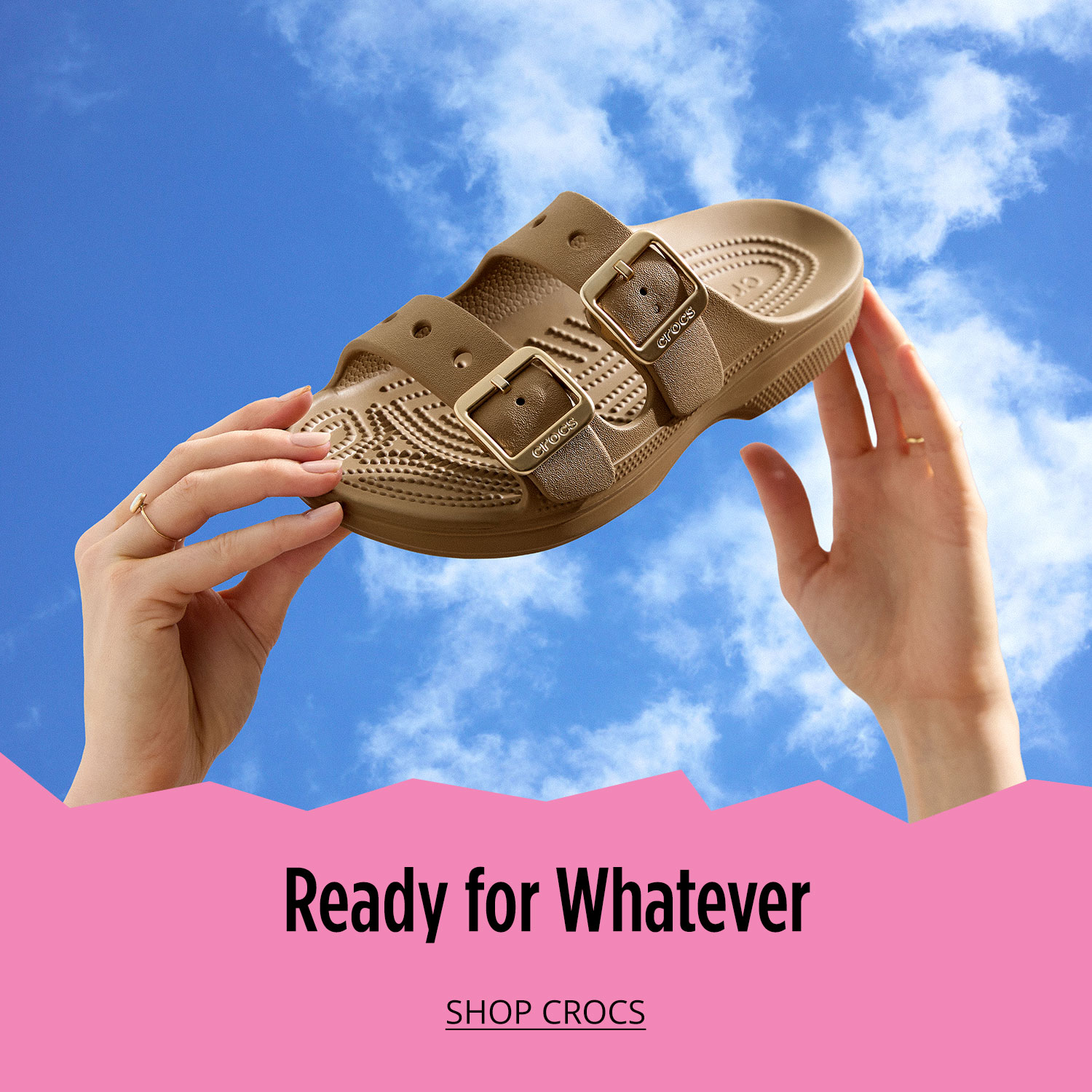 Two hands holding a pair of Crocs. Background is a blue sky with clouds  
