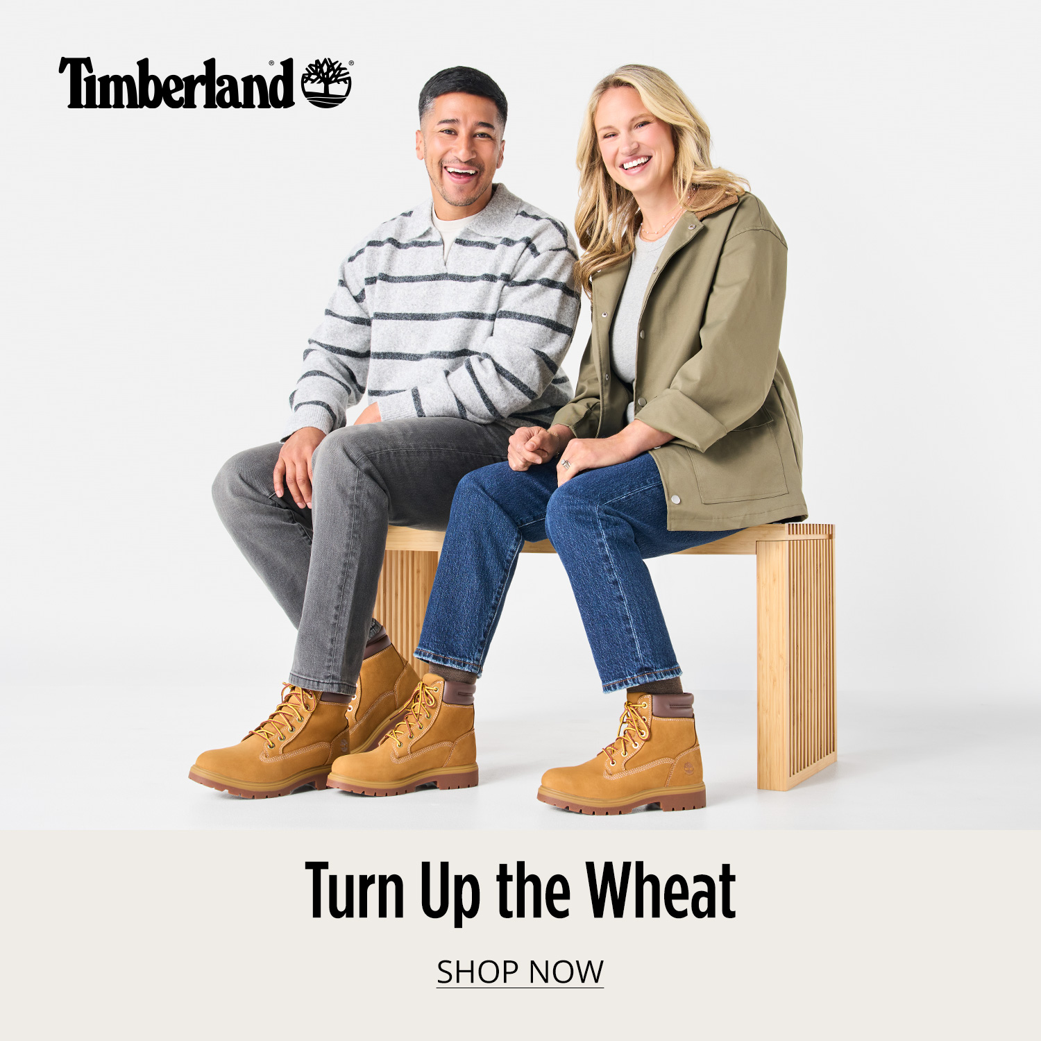 A stylish man and woman sitting on a bench dressed casually and both wearing Timberland wheat lace-up boots with text saying Turn Up the Wheat