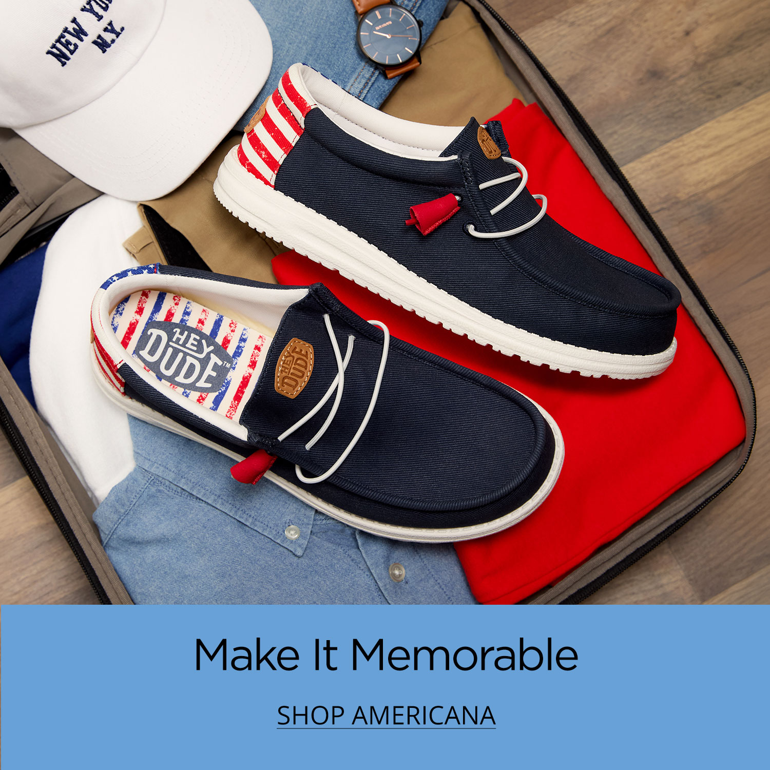 A pair of all-American, red, white and blue HEYDUDE shoes sitting inside a packed suitcase on a wooden floor.