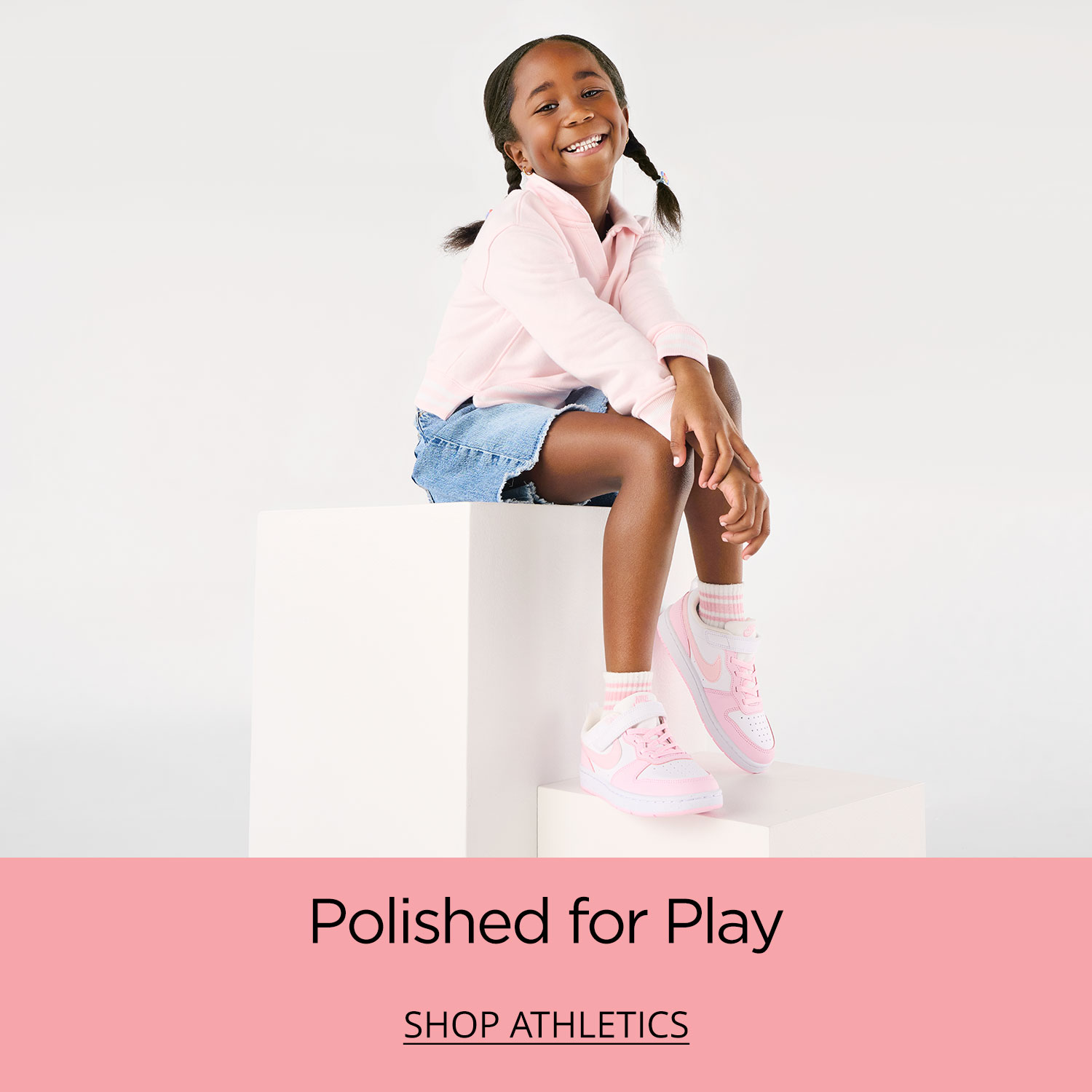 A young girl sitting on a plinth ready to jump into play with her pink and white Nike Court Borough sneakers.