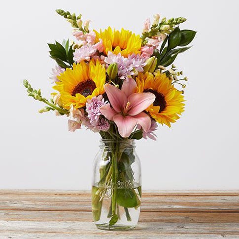 A bouquet of sunflowers, lilies, and snapdragons arranged in a clear glass vase on a wooden surface.