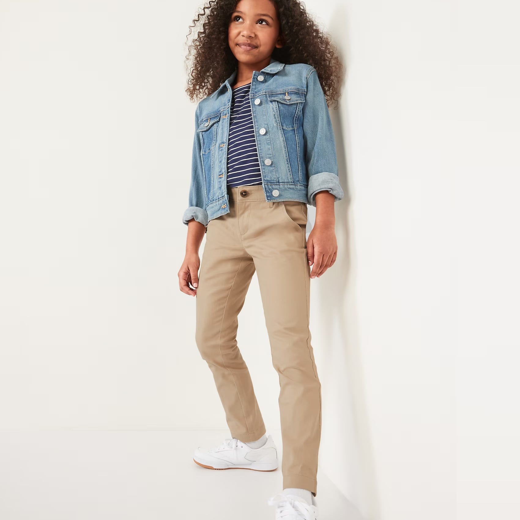 A young girl models a denim jacket, striped top, khaki pants, and white sneakers.