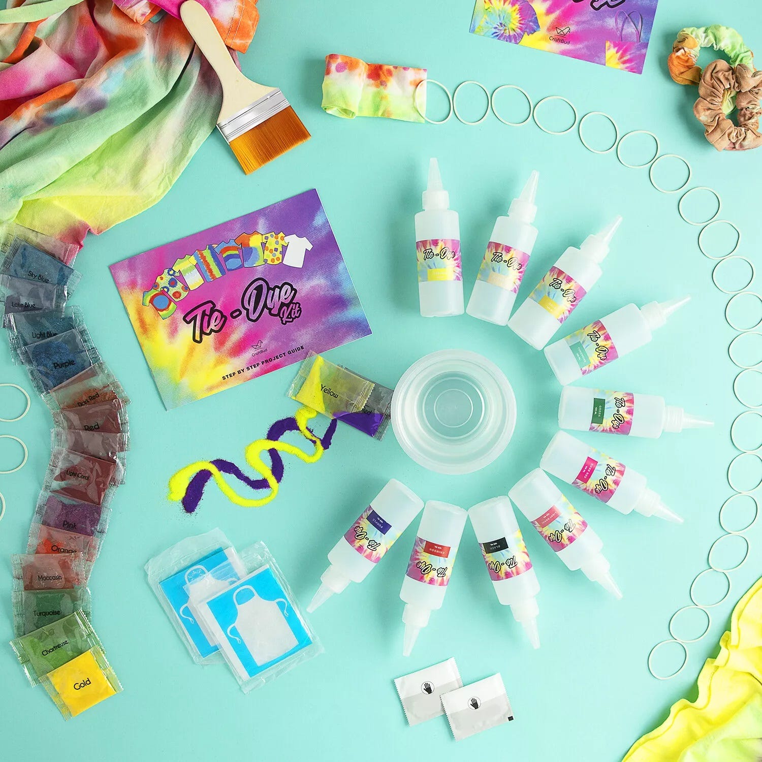 A tie-dye kit with various colored dyes in squeeze bottles, fabric items, rubber bands, and protective gloves on a teal background.