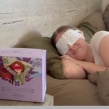 A person lies on a bed wearing a scented eye mask, with a box labeled \“Frudia Scented Steam Eye Mask\“ nearby, featuring an illustration of a relaxed person with an eye mask.