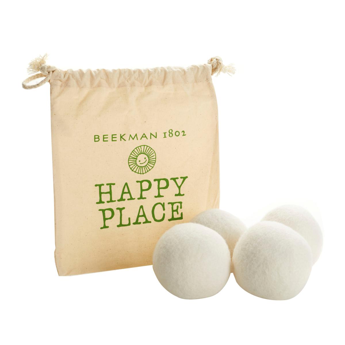 A set of four white wool dryer balls is shown alongside a beige drawstring bag labeled \“Beekman 1802 Happy Place,\“ designed to reduce drying time and soften laundry.