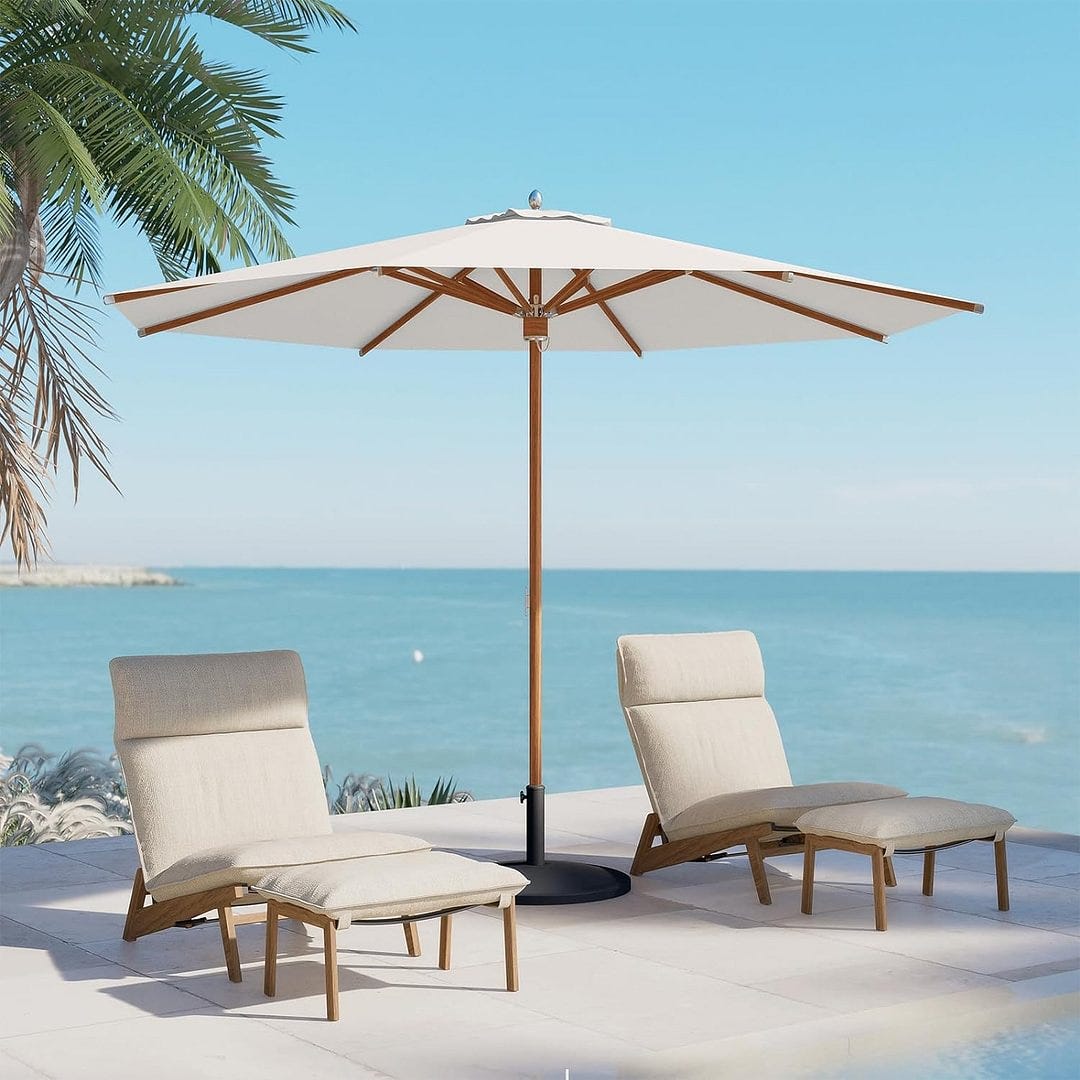 A large outdoor umbrella with a wooden pole and two cushioned lounge chairs by a pool facing the sea.