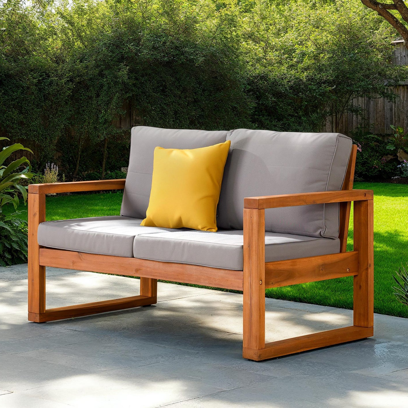 Outdoor wooden loveseat with gray cushions and a yellow throw pillow, set on a stone patio with a lush green yard in the background.