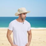 A man on a beach wears a beige wide-brim sun hat with UPF 50 protection and an adjustable chin strap.
