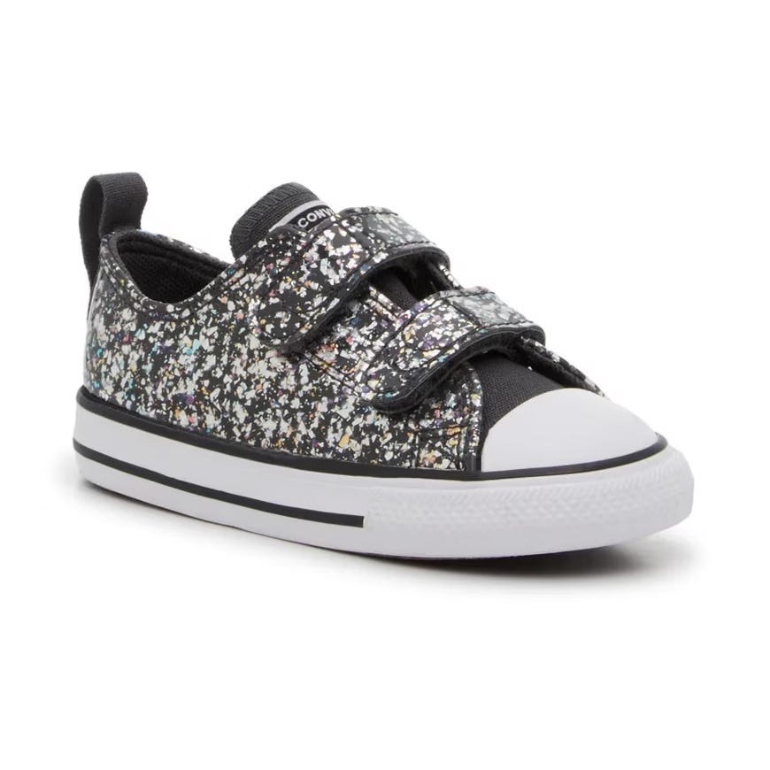 A child's black sneaker with multicolored speckles, featuring a white toe cap and two Velcro straps.
