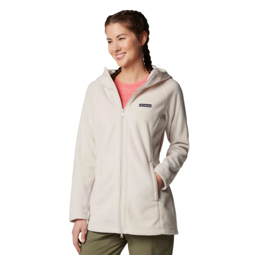 A woman is wearing a light-colored hooded fleece jacket with a front zipper and pockets, paired with a red shirt underneath and green pants.