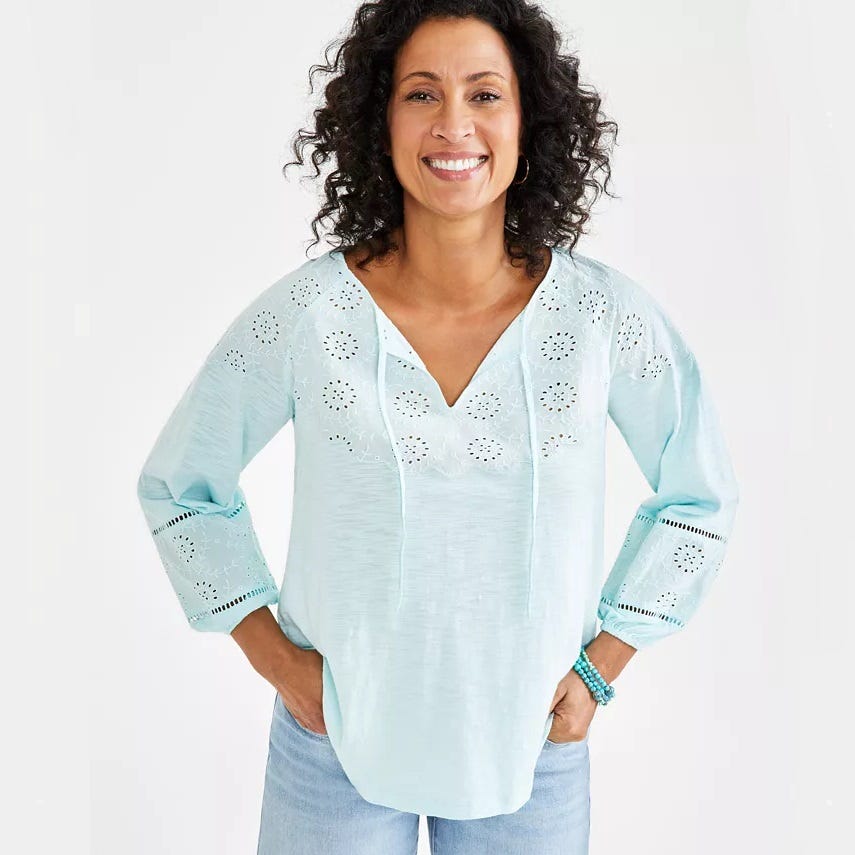 A woman wearing a light blue tunic with decorative eyelet detailing and rolled-up ¾ sleeves.