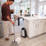 A man sweeps debris into a stationary vacuum device in a kitchen while a woman works at a table in the background.